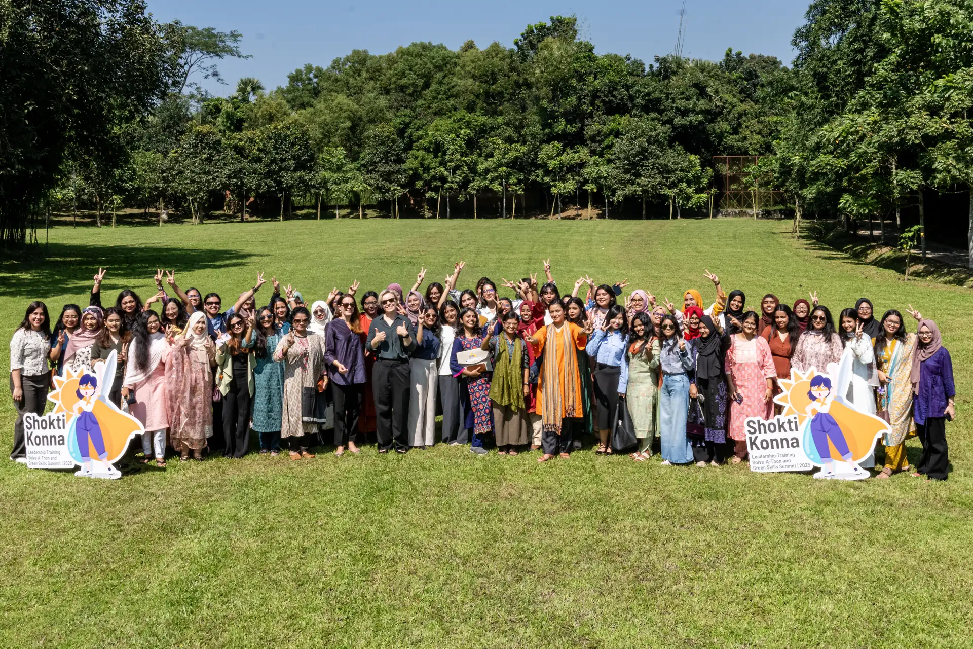Shoktikonna cohort members flashing victory signs on a lawn beside Shoktikonna standees